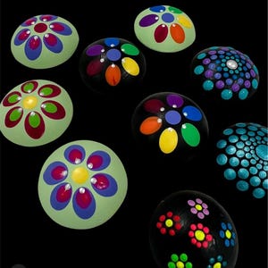 Può includere: Una collezione di otto pietre rotonde dipinte con disegni colorati. Alcune pietre presentano grandi motivi a petali multicolori con punti centrali, mentre altre mostrano densi arrangiamenti di piccoli punti dipinti dai colori vivaci.