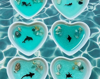 May include: Six heart-shaped dishes filled with turquoise blue resin, each containing a different sea creature silhouette, seashells, and small white stones.