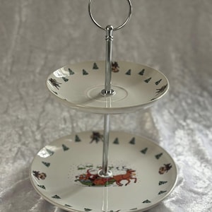 Könnte beinhalten: Zweistöckiger Servierständer mit silbernem Griff. Die weißen Keramikplatten weisen ein festliches Design mit Hirschen, Bäumen und anderen festlichen Elementen auf. Ideal zum Servieren von Desserts oder Vorspeisen.