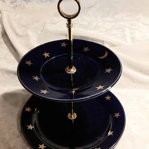 Könnte beinhalten: Zweistöckiger Servierständer mit marineblauen Tellern, verziert mit goldenen Sternen und einem Mond. Der Ständer hat einen goldfarbenen Griff und eine zentrale Stütze. Ein dekoratives Stück zum Servieren von Desserts oder Vorspeisen.