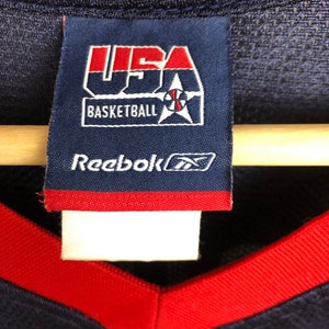 May include: A blue and red Reebok basketball jersey with a USA Basketball logo and Reebok logo tag. The tag has the text "USA Basketball" and "Reebok" in white.