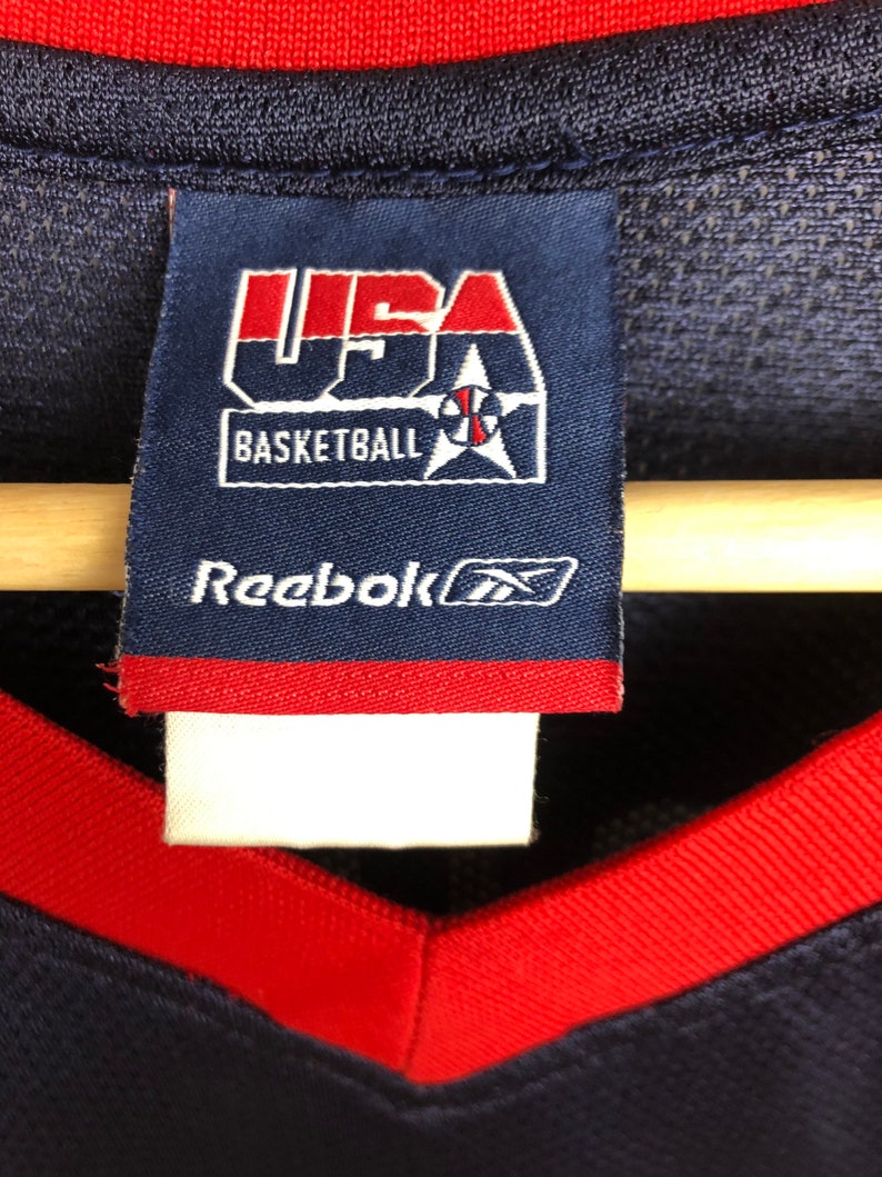 May include: A blue and red Reebok basketball jersey with a USA Basketball logo and Reebok logo tag. The tag has the text "USA Basketball" and "Reebok" in white.