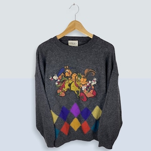 May include: A grey sweater featuring a colourful cartoon design of a horse-drawn carriage with multiple characters. The sweater has a diamond pattern at the bottom in shades of yellow, orange, red, purple and blue.