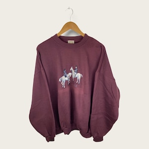 May include: A maroon sweatshirt with an embroidered design of two people riding horses.