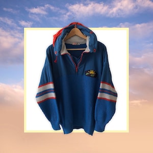 May include: A blue and red hooded sweater with white stripes on the sleeves. The sweater has a zipper and a patch with a sun and mountains on the front. The sweater is made of a soft, knitted fabric.