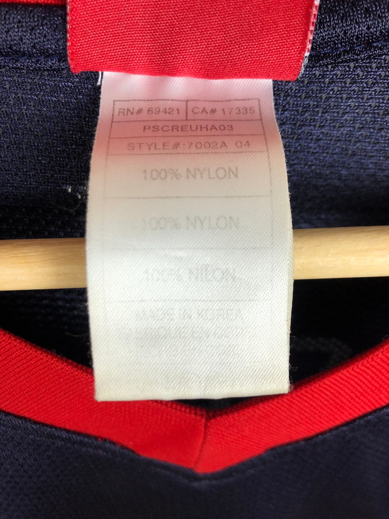 May include: A white tag with black text that reads "RN# 69421 CA# 17335 PSCREUHA03 STYLE#:7002A 04 100% NYLON 100% NYLON".