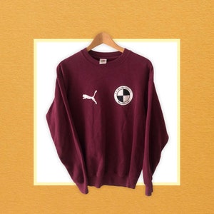 May include: A maroon long-sleeved sweatshirt with a white Puma logo on the left chest and a white circular logo with the text "AS Bicha 1911" and a black and white chequered design on the right chest.