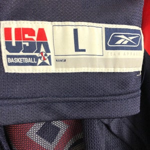 May include: A navy blue basketball jersey with a white tag that says "USA Basketball" with a basketball and star logo. The tag also says "L" for size and "Team Apparel" with a Reebok logo.