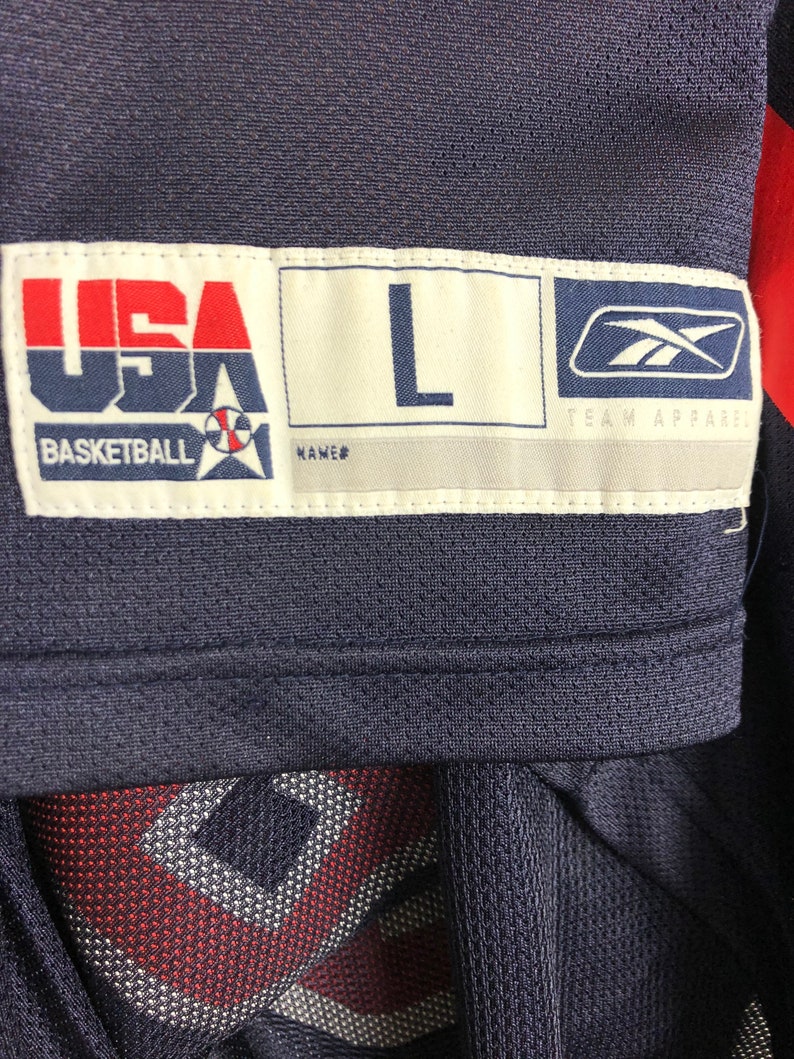 May include: A navy blue basketball jersey with a white tag that says "USA Basketball" with a basketball and star logo. The tag also says "L" for size and "Team Apparel" with a Reebok logo.