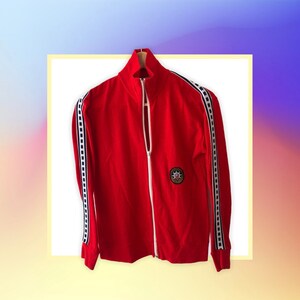 May include: Red zippered jacket with black and white striped trim on the sleeves and a circular patch on the chest.