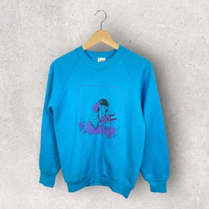 May include: A turquoise blue sweatshirt featuring a graphic of a girl holding a bird. The graphic is framed with a pink dotted line and surrounded by pink flowers.