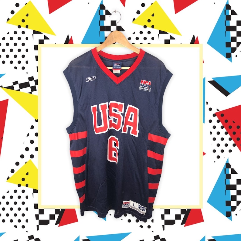 May include: A navy blue basketball jersey with red and white stripes. The jersey has "USA" printed on the front in red and white lettering with the number "6" underneath. The jersey is sleeveless and has a V-neck.