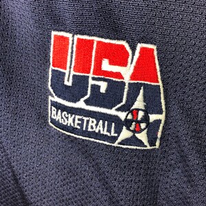 May include: A navy blue basketball jersey with a red, white and blue embroidered patch that says "USA Basketball".