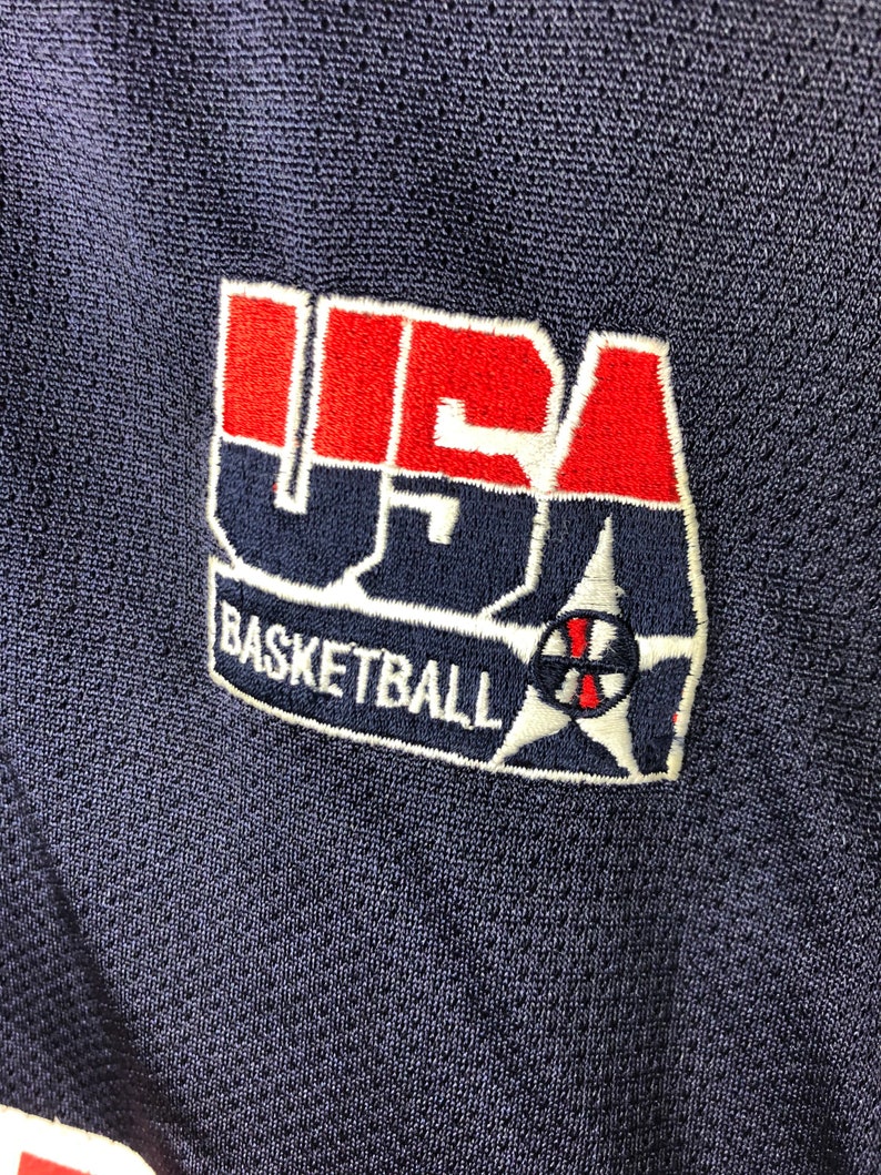 May include: A navy blue basketball jersey with a red, white and blue embroidered patch that says "USA Basketball".