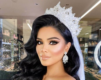 May include: A woman with long, wavy black hair, wearing a sparkling silver crown and matching earrings. She is dressed in a black off-the-shoulder top, with a sheer veil. The background is a mirrored wall with shelves.