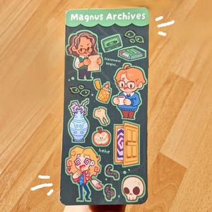 以下が含まれることがあります： 上部に「Magnus Archives」の文字が記された長方形のしおり。ダークグリーンの表面には、キャラクター、カセットテープ、ライター、花瓶、歯、リンゴ、ドア、頭蓋骨など、様々な漫画風のイラストが描かれています。