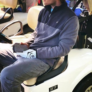 Peut inclure: Une personne portant un pull &agrave; col roul&eacute; gris et noir, des gants de golf blancs et un jean bleu est assise dans une voiturette de golf blanche avec le num&eacute;ro 27 sur le c&ocirc;t&eacute;. La voiturette a un si&egrave;ge noir et un volant noir.