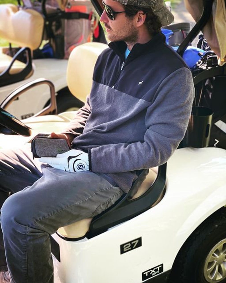Peut inclure: Une personne portant un pull &agrave; col roul&eacute; gris et noir, des gants de golf blancs et un jean bleu est assise dans une voiturette de golf blanche avec le num&eacute;ro 27 sur le c&ocirc;t&eacute;. La voiturette a un si&egrave;ge noir et un volant noir.