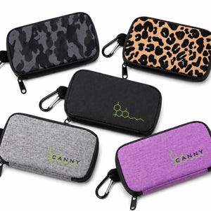 May include: Five rectangular zippered cases with carabiner clips. One features a dark gray camouflage pattern, one has a tan and black leopard print, one is solid black with a green chemical structure, one is heather gray with the text CANNY, and one is purple with the text CANNY.