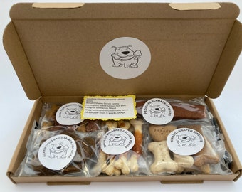 May include: An open cardboard box filled with various dog treats. The box contains six individual bags of treats, each with a different brand and type of treat. The bags are labeled with the names of the treats, such as "Wag Tasties Chicken & Liver", "Harringtons Baked Salmon Fish Bites", "Chicken Wrapped Calcium Bones", "Pedigree Schmackos Mixed", "Winalot Shaped Biscuits", and "Shaftesbury Biscuit Variety".