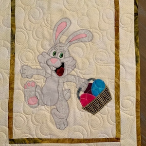 May include: A gray bunny rabbit with pink ears is holding a basket of colorful Easter eggs. The bunny is running and has a happy expression on its face. The background is a white quilted fabric with a brown and gold border.