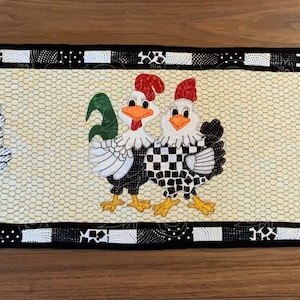 Może przedstawiać: Czarno-biały bieżnik kuchenny w kratkę z czarną obwódką i białym tłem. Na bieżniku wyszyte są trzy kolorowe kury. Kury są czarne, białe i czerwone z zielonymi i różowymi akcentami.