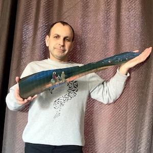 Könnte beinhalten: Ein langes, rechteckiges Holzkunstwerk mit einer gemalten Landschaft mit Figuren zu Pferd. Das Kunstwerk hat einen dunkelblauen und orangefarbenen Himmel, mit Figuren im Vordergrund. Das Stück wird von einer Person gehalten, die einen grauen Pullover trägt.