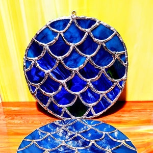 May include: Two round, stained glass pieces with a mermaid scale design. The glass is a deep blue with silver-colored metal edging. One piece is suspended, and the other rests below on a wooden surface.