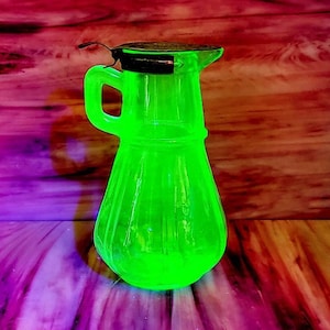 May include: A vibrant, neon green glass pitcher with a handle and a black lid. The pitcher has a ribbed design and a spout for pouring. The background features a colorful, abstract pattern of pink, purple, and brown.
