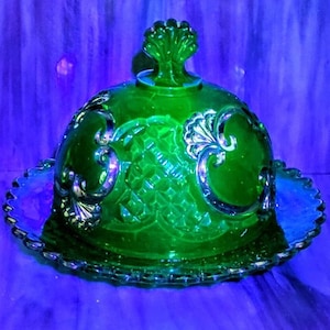 May include: A green glass covered dish with a decorative lid and a matching base. The dish has a detailed, embossed pattern and a scalloped edge. The glass has a vibrant green color and a glossy finish.