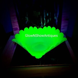 May include: A glowing green glass bowl with a scalloped edge. The bowl is square-shaped with a slightly curved interior. The text "GlowNShowAntiques" is visible on the bowl. The background is dark.
