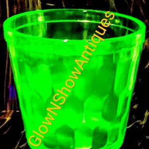 Puede incluir: Un vaso de vidrio verde brillante, de estilo vintage. El vaso tiene una superficie ligeramente texturizada y un borde acampanado. Las palabras "GlowNShowAntiques" están escritas en diagonal sobre el vaso en amarillo-naranja. El fondo es oscuro y borroso.