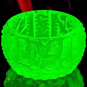 May include: A glowing green glass bowl with a detailed floral pattern. The bowl has a rounded shape and sits on a reflective surface, creating a mirrored effect. The glass appears to be vintage or antique.