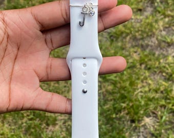 Pode incluir: Pulseira branca para Apple Watch com um berloque decorativo. O berloque inclui um pingente de letra "J" prateado, uma pequena gema transparente e contas rosa. A pulseira é segurada contra um fundo de grama verde.