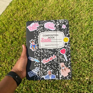 May include: Black composition book style tablet case with various stickers, including a pink cowboy hat, a smiley face, and the text "Jade". The case is designed by Fintle in the USA. Other stickers include "anxiety Prime" and "Thick-fil-a".