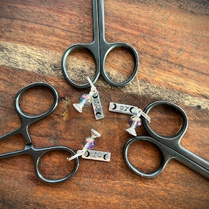 May include: Three silver charm pendants with a moon and star design, each attached to a small glass vial with iridescent purple and blue beads. The charms are arranged on a wooden surface with black surgical scissors.