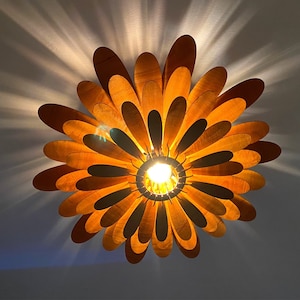 May include: A wooden ceiling light fixture shaped like a flower with multiple layers of petals. The petals are a light brown colour and the light fixture is illuminated by a single light bulb in the centre.