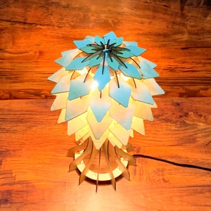 Op de afbeelding: Een teal en witte houten lamp met een geometrisch ontwerp. De lamp is aan en geeft een warm licht.