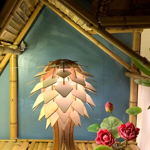Peut inclure: Une lampe en bois au design original, avec un abat-jour en forme de p&eacute;tales superpos&eacute;s. La lampe est pos&eacute;e sur une surface en bambou, avec des fleurs et des feuilles de lotus artificielles. L'arri&egrave;re-plan comprend des structures en bambou et un mur turquoise.