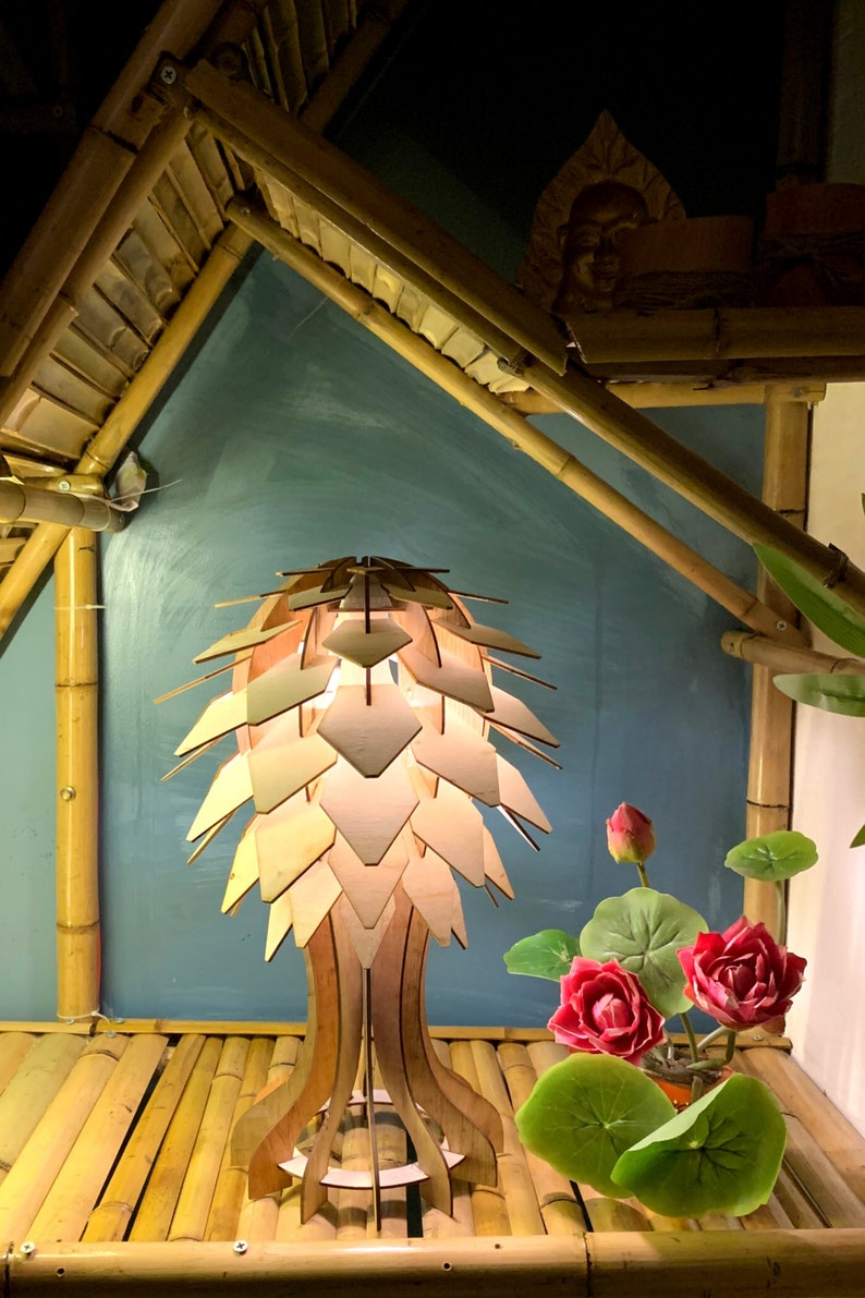 Peut inclure: Une lampe en bois au design original, avec un abat-jour en forme de p&eacute;tales superpos&eacute;s. La lampe est pos&eacute;e sur une surface en bambou, avec des fleurs et des feuilles de lotus artificielles. L'arri&egrave;re-plan comprend des structures en bambou et un mur turquoise.
