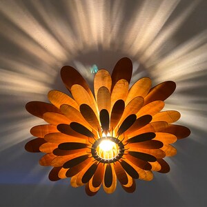 May include: A wooden ceiling light fixture shaped like a flower with multiple layers of petals. The light is on and casts a warm glow.