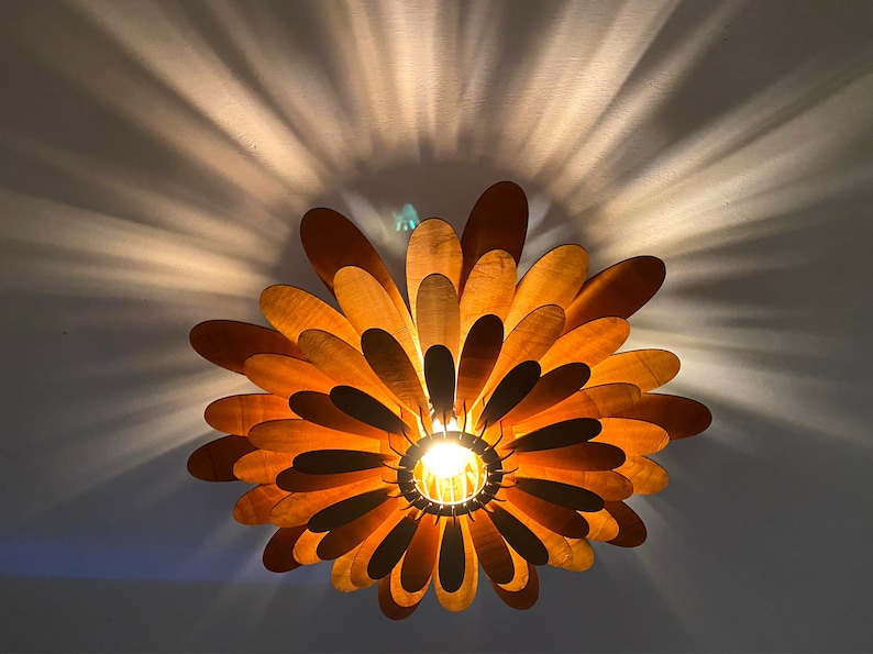 May include: A wooden ceiling light fixture shaped like a flower with multiple layers of petals. The light is on and casts a warm glow.