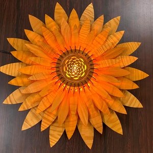Puede incluir: Una l&aacute;mpara de madera con forma de girasol con un acabado marr&oacute;n claro y un brillo amarillo c&aacute;lido. La l&aacute;mpara tiene un centro circular con un dise&ntilde;o geom&eacute;trico.