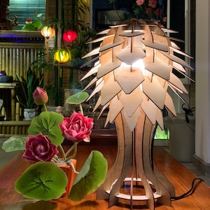 Peut inclure: Lampe de table en bois au design unique, avec un abat-jour clair en couches et une base en bois fonc&eacute;. La lampe est plac&eacute;e &agrave; c&ocirc;t&eacute; d'un petit pot orange avec des fleurs et des feuilles de lotus artificielles. L'arri&egrave;re-plan comprend des lanternes color&eacute;es et une fen&ecirc;tre.