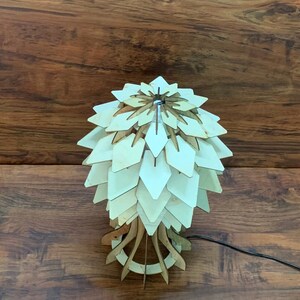 Peut inclure: Une lampe de table en bois au design unique. La lampe pr&eacute;sente un abat-jour en forme de p&eacute;tales superpos&eacute;s dans des tons de bois clair, soutenu par une base g&eacute;om&eacute;trique. La lampe est reli&eacute;e &agrave; un cordon d'alimentation noir. Le fond est une surface en bois fonc&eacute;.