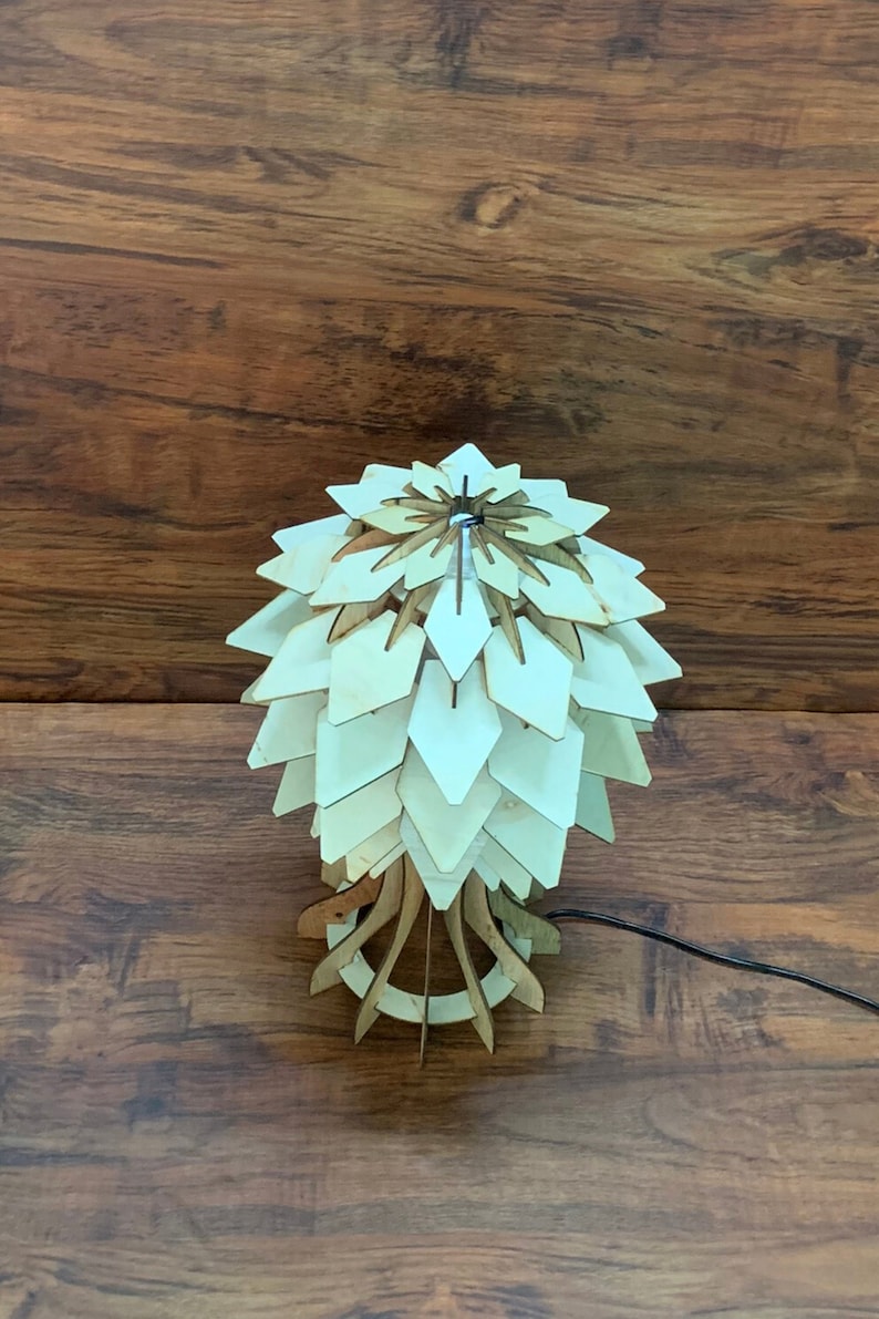 Peut inclure: Une lampe de table en bois au design unique. La lampe pr&eacute;sente un abat-jour en forme de p&eacute;tales superpos&eacute;s dans des tons de bois clair, soutenu par une base g&eacute;om&eacute;trique. La lampe est reli&eacute;e &agrave; un cordon d'alimentation noir. Le fond est une surface en bois fonc&eacute;.