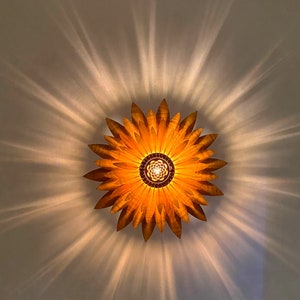 Puede incluir: Una l&aacute;mpara de madera con forma de girasol con un c&aacute;lido brillo dorado. La l&aacute;mpara proyecta un patr&oacute;n de estrella en el techo.