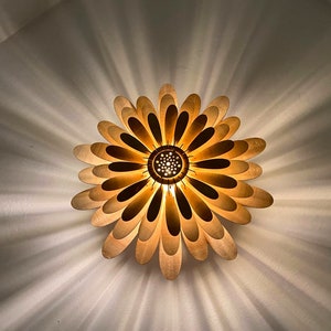 May include: A wooden ceiling light fixture shaped like a flower with multiple petals. The light is on and illuminates the room with a warm glow.