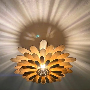 May include: A wooden ceiling light fixture shaped like a flower with multiple layers of petals. The light is on and casts a warm glow.