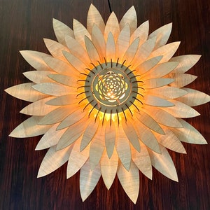 Op de afbeelding: Een houten lamp in de vorm van een zonnebloem met een gloeilamp in het midden. De lamp heeft een natuurlijke houten afwerking en wordt verlicht door een warm licht.